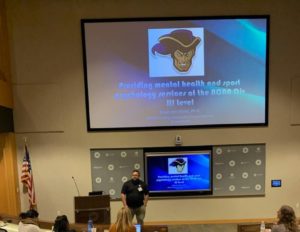 Dr. Jason von Stietz stands near a podium and a giant flat screen and discusses his work providing mental health and sport psychology services for NCAA athletes